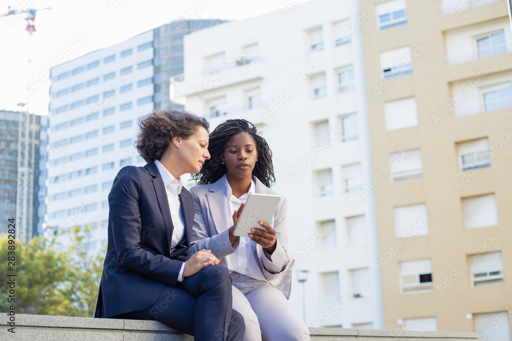 Serious female colleagues with tablet pc. Low angle view of multiethnic young businesswomen sitting together and using digital tablet outdoor. Technology concept Serious female colleagues with tablet pc. Low angle view of multiethnic young businesswomen sitting together and using digital tablet outdoor. Technology concept