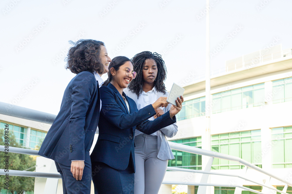 Cheerful businesswomen with tablet computer. Smiling multiethnic female colleagues standing and using digital tablet together outdoor. Technology concept Cheerful businesswomen with tablet computer. Smiling multiethnic female colleagues standing and using digital tablet together outdoor. Technology concept