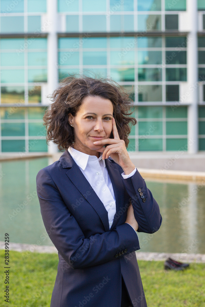 Pensive satisfied businesswoman posing outside. Woman wearing formal suit, standing near office building, touching face and smiling at camera. Confident business lady concept Pensive satisfied businesswoman posing outside. Woman wearing formal suit, standing near office building, touching face and smiling at camera. Confident business lady concept