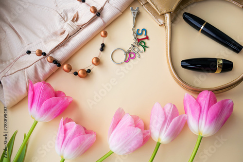 A composition of five buds of pink flowers, fragments of a handbag, cosmetics, dress, necklace and keychain similar to the Eiffel Tower with the inscription Paris. Top view on a beige background