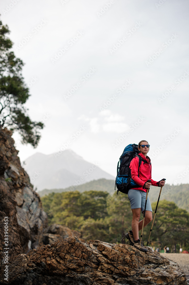 Slim fit girl in sportwear standing with hiking backpack and sticks on the rock