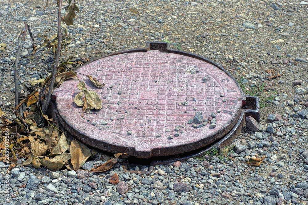 brown iron manhole in dry leaves on the gray sand of the road brown iron manhole in dry leaves on the gray sand of the road