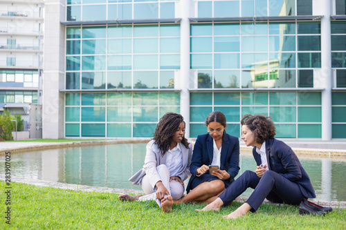 Barefoot multiethnic businesswomen with smartphone outdoor. Young female colleagues sitting on green lawn and using mobile phone. Technology concept © Mangostar
