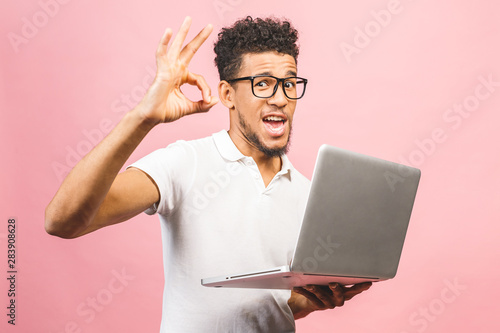 Foto Portrait of a happy young african man in casual holding screen laptop computer while showing ok sign gesture and looking at camera isolated over pink background