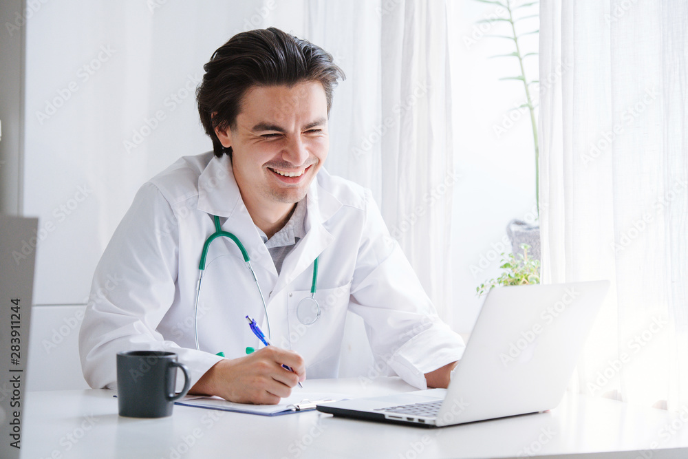 Male doctor working with laptop behind desk, writing on paperwork in a hospital. Male doctor working with laptop behind desk, writing on paperwork in a hospital.