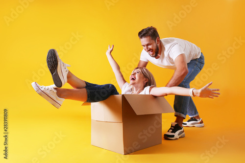 Young married couple moving into a new home. Attractive blonde woman sitting in cardboard box while bearded man pushes her. Newely weds fooling around. Isolated yellow background, copy space, close up