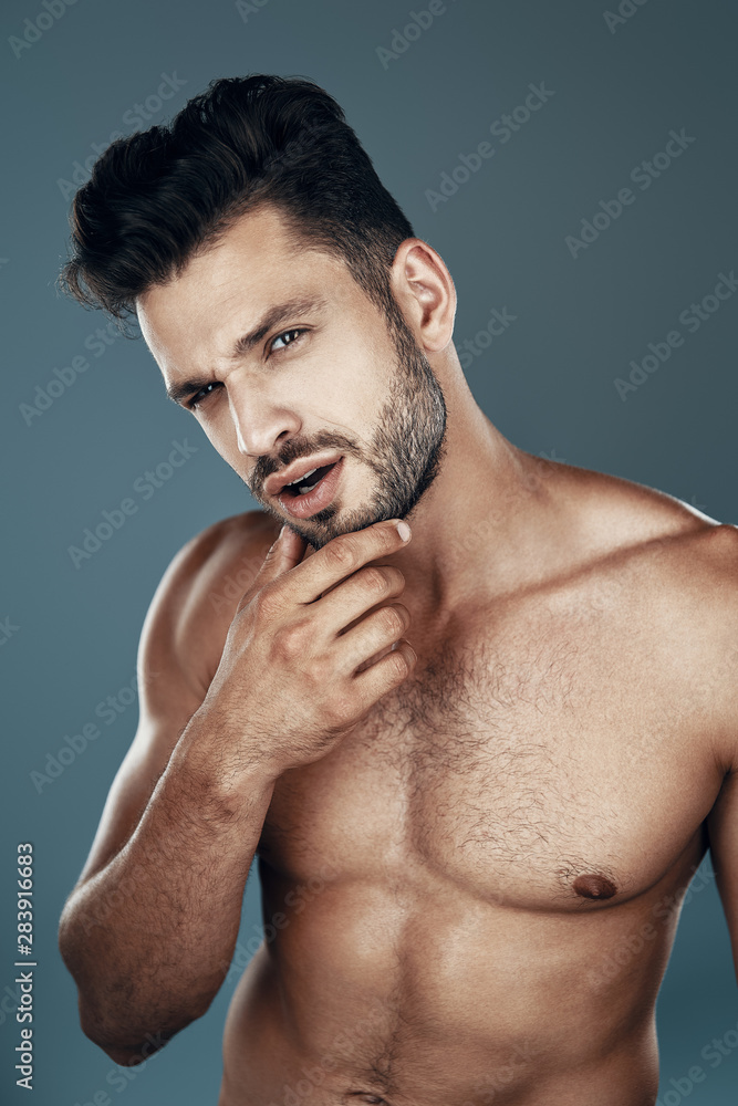 Obraz premium Handsome young shirtless man looking at camera and keeping hand on chin while standing against grey background