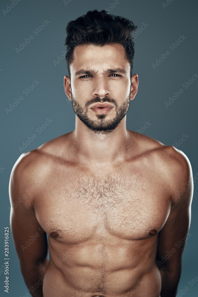Handsome young shirtless man looking at camera while standing against grey background Handsome young shirtless man looking at camera while standing against grey background