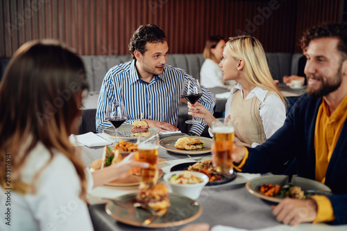 Multiethnic friends sitting at restaurant, drinking alcohol, chatting and having burgers for dinner.