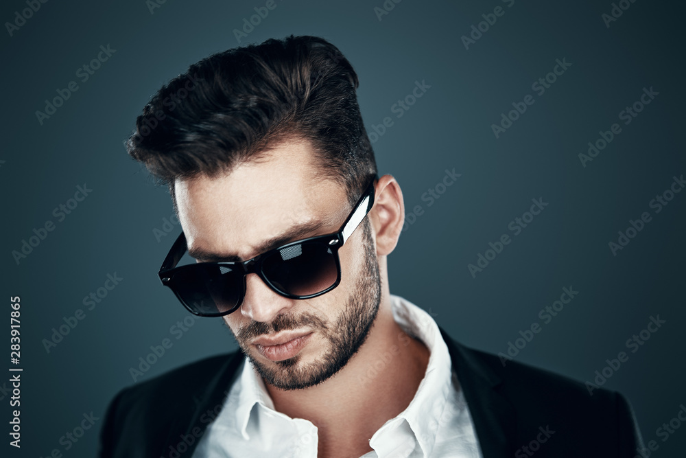 Charming young man in eyewear looking down while standing against grey background Charming young man in eyewear looking down while standing against grey background