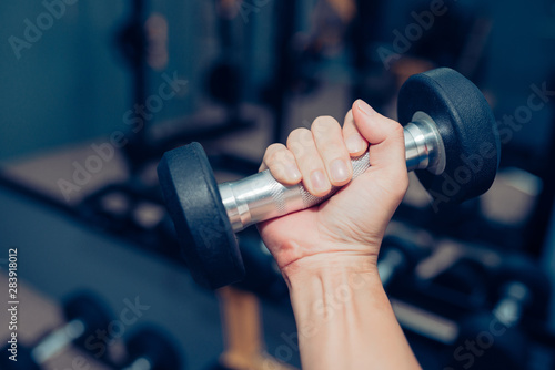 Hands are holding a black dumbbell in the gym with a backdrop of fitness room and sun light.