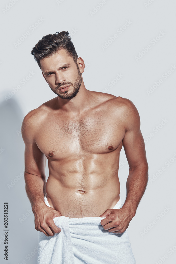 Fototapeta premium Handsome young shirtless man looking at camera while standing against grey background