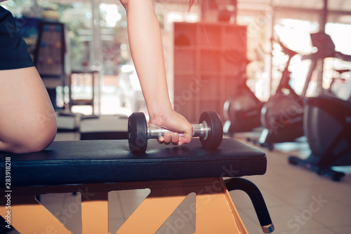 Hands are holding a black dumbbell in the gym with a backdrop of fitness room and sun light.