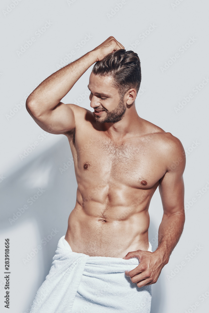 Handsome young shirtless man covered with towel smiling while standing against grey background