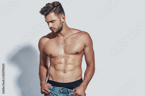 Handsome young shirtless man taking off his jeans while standing against grey background