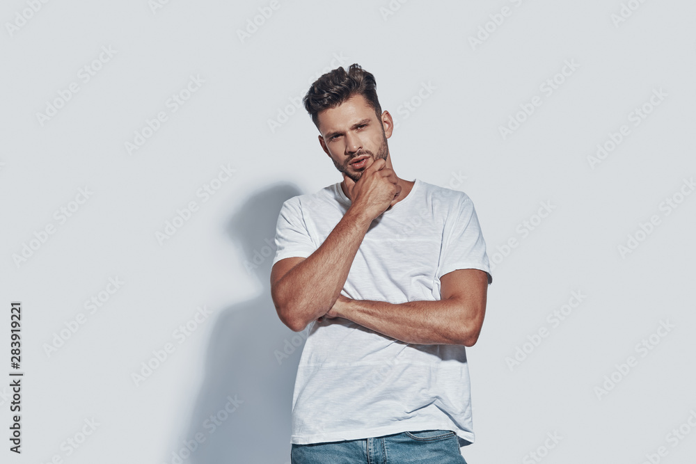 Handsome young man looking at camera and keeping hand on chin while standing against grey background