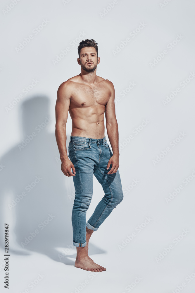 Fototapeta premium Full length of handsome young shirtless man looking at camera while standing against grey background