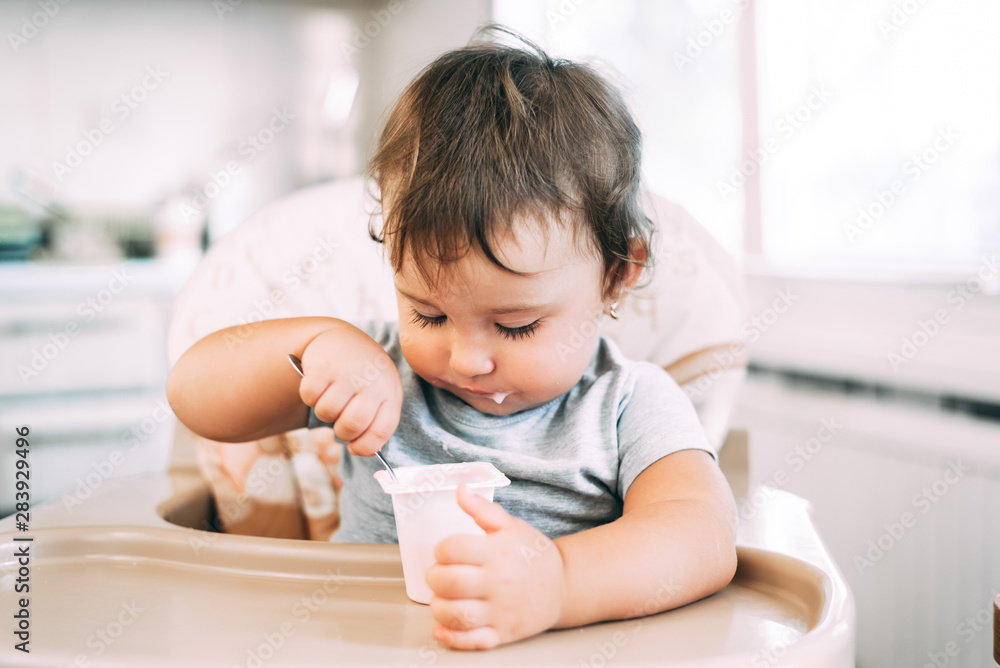 Cute, little girl eating yogurt in the kitchen during the day in a high chair Cute, little girl eating yogurt in the kitchen during the day in a high chair