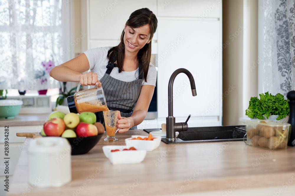 Beautiful smiling Caucasian woman in apron standing in kitchen and pouring fresh smoothie into glass. Beautiful smiling Caucasian woman in apron standing in kitchen and pouring fresh smoothie into glass.