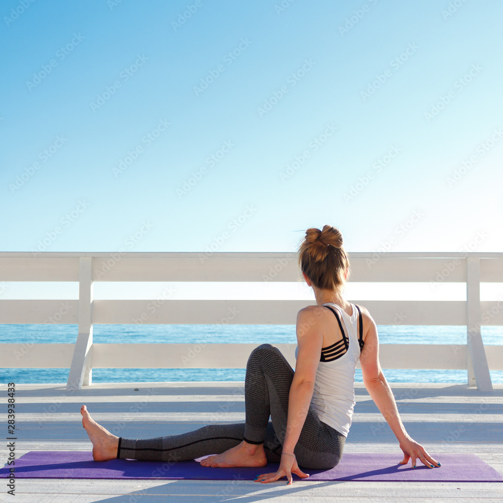 Young slim woman in tight sportswear practicing yoga outdoors at white wooden seafront. Sitting twisting pose.