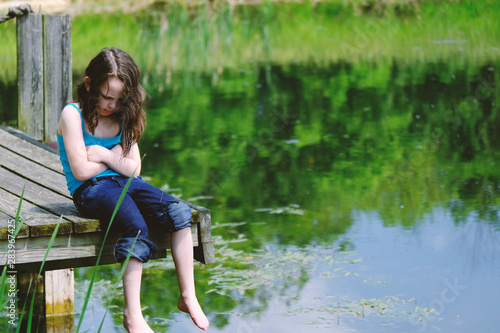 Fototapeta Naklejka Na Ścianę i Meble -  Young girl sitting on dock by rural pond water, arms crossed.  Concept of upset child.