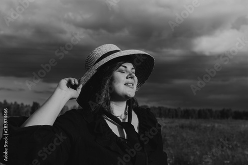 Attractive curvy woman in a straw hat on the background of a stormy sky. Stylish accessories, seashells, straw hat, fashion for plus size. Body positive, natural authentic beauty. Black and white. © Julia