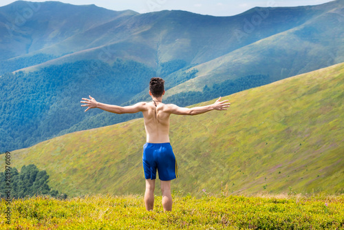 A young man is resting in the mountains in the summer and feels complete freedom and personality growth outdoors. The guy stands with his back to the camera and stretches out his hands