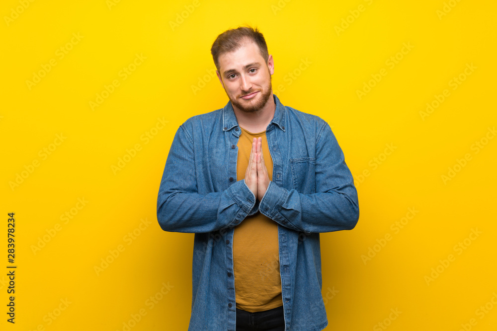 Blonde man over isolated yellow wall keeps palm together. Person asks for something Blonde man over isolated yellow wall keeps palm together. Person asks for something