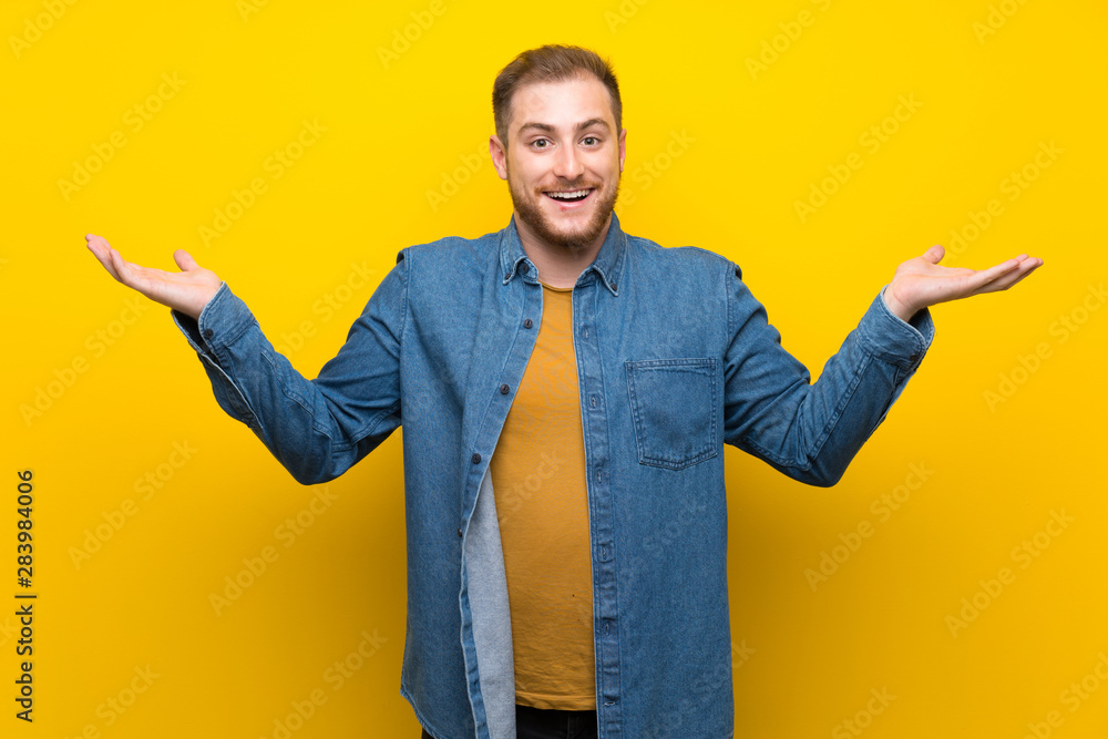 Blonde man over isolated yellow wall with shocked facial expression Blonde man over isolated yellow wall with shocked facial expression