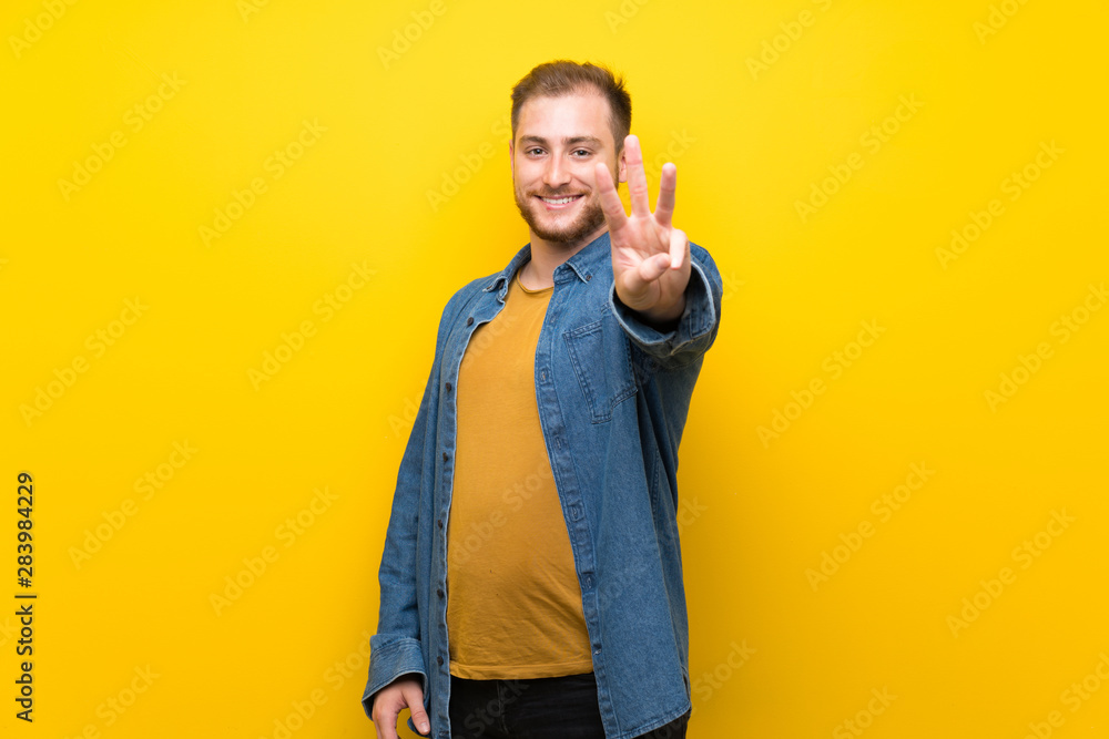 Blonde man over isolated yellow wall happy and counting three with fingers Blonde man over isolated yellow wall happy and counting three with fingers