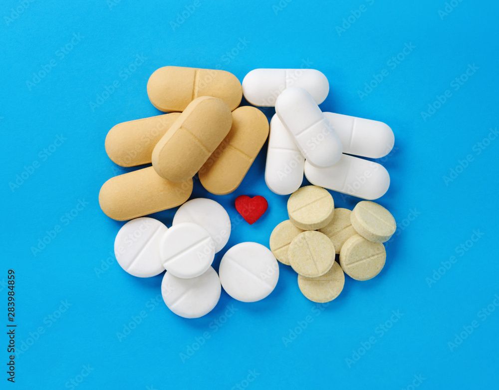 Different tablets, pills and small red heart in center on blue background. Top view. Medical concept. Different tablets, pills and small red heart in center on blue background. Top view. Medical concept.