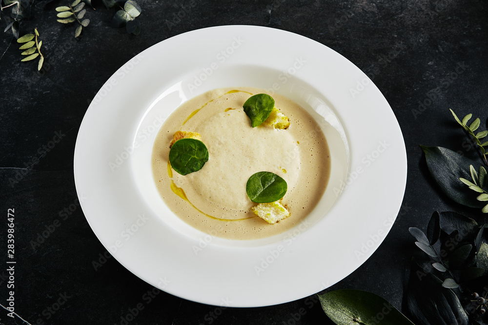 Exquisite Serving White Restaurant Plate of Cream Cappuccino Soup with Porcini Mushrooms and Truffle Close Up. Beautiful Creative Molecular Dish with Boletus on Black Stone and Plants Background