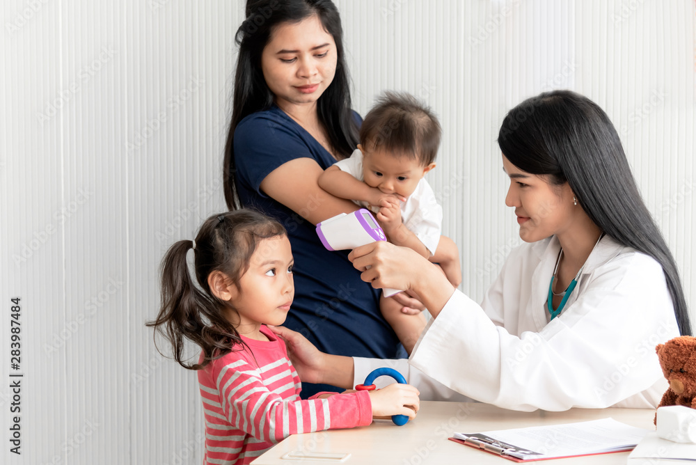 Asian woman doctor using Infraed Thermometer with a girl, whose mother took the son and daughter to receive treatment from the flu, Which is pandemic, to health care and health insurance concept. Asian woman doctor using Infraed Thermometer with a girl, whose mother took the son and daughter to receive treatment from the flu, Which is pandemic, to health care and health insurance concept.