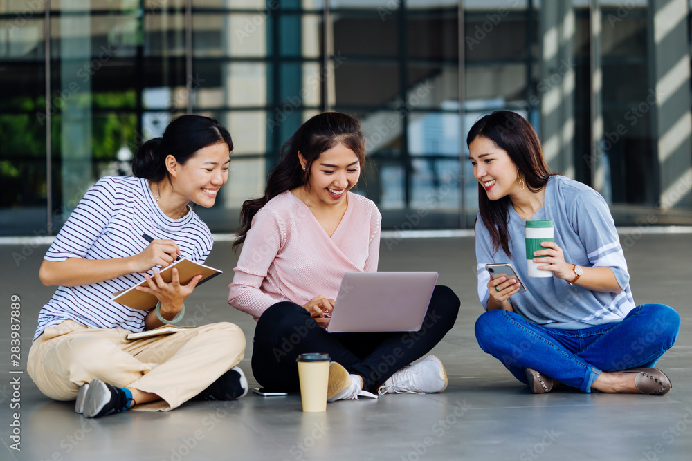 Group of young smart Asian students laughing making notes looking at laptop and discussing while sitting on floor outdoors Group of young smart Asian students laughing making notes looking at laptop and discussing while sitting on floor outdoors