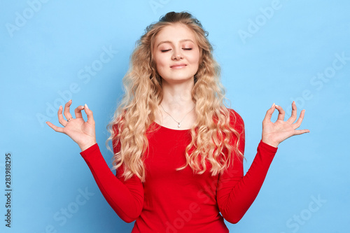 relaxed blonde girl dressed in red sweater keeping eyes closed while mmeditating indoors, practicing peace of mind, keeping finges in mudra gesture, hobby, interest, lifestyle, free time, spare time photo