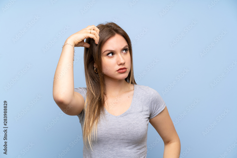 Teenager girl over isolated blue wall having doubts while scratching head Teenager girl over isolated blue wall having doubts while scratching head