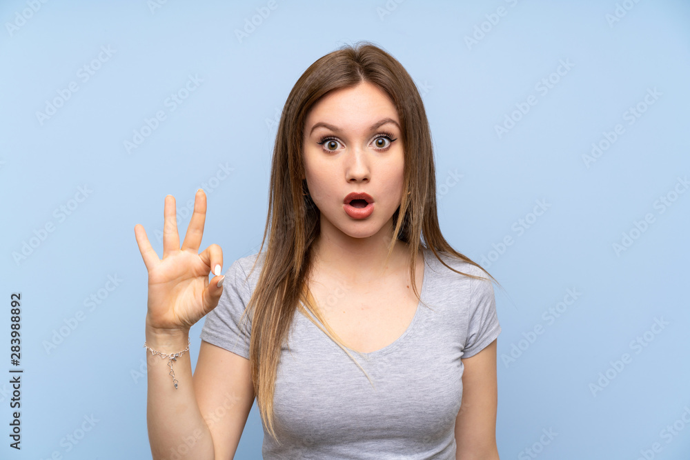 Teenager girl over isolated blue wall surprised and showing ok sign Teenager girl over isolated blue wall surprised and showing ok sign