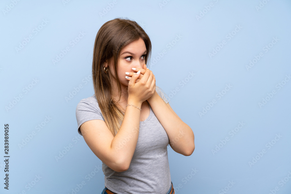 Teenager girl over isolated blue wall covering mouth and looking to the side Teenager girl over isolated blue wall covering mouth and looking to the side