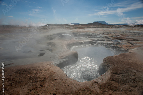 A wide angle view of icelandic volcanic landscape.Mud pot bubbling in foreground and steam vents in distance with volcanic mountains on horizon - Image