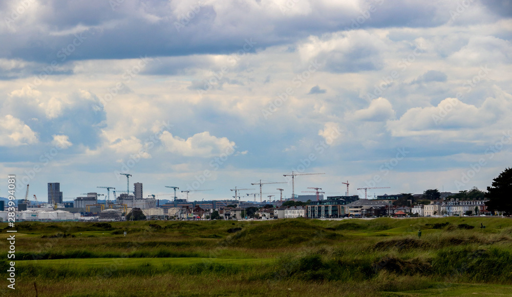 Cranes along the Dublin skyline with the green grass of Bull Island causeway in the foreground and blue sky and white fluffy clouds above the city scene.