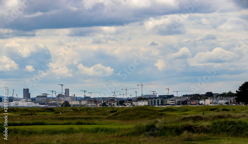 Cranes along the Dublin skyline with the green grass of Bull Island causeway in the foreground and blue sky and white fluffy clouds above the city scene.