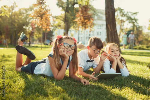 Three smiling school-age friends are lying on the grass and looking at the camera. In the hands of children a tablet