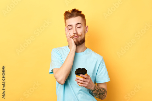 Tired exhausted guy with sleepy expression, covers face with palm, holds disposable paper cup of coffee. isolated yellow background