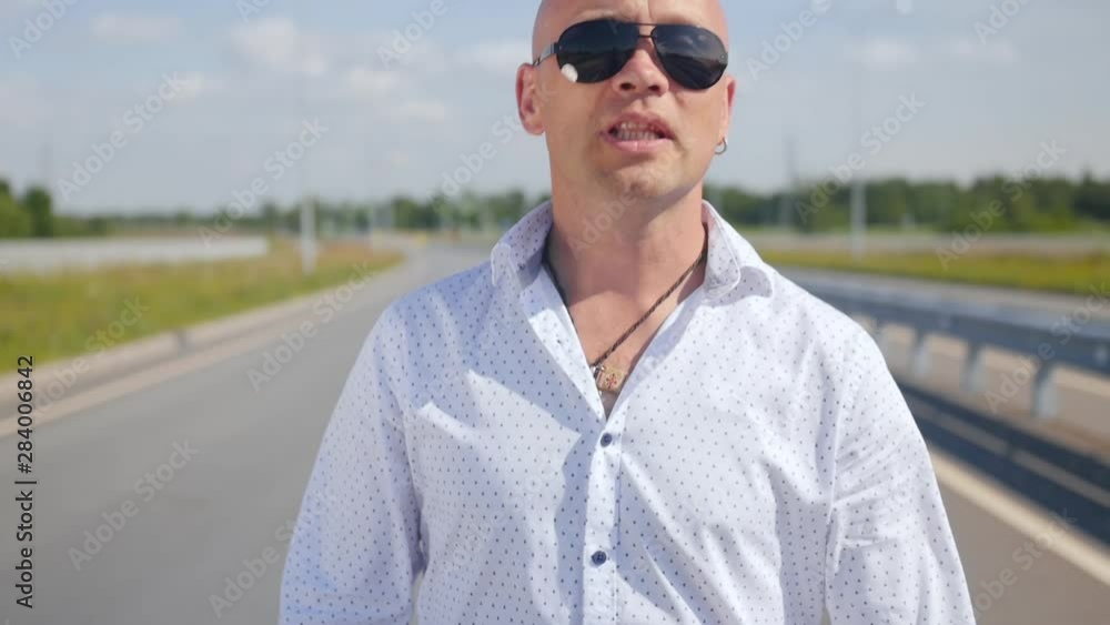 Portrait of a bald man in sunglasses singing and walking along the road. Medium shot. Portrait of a bald man in sunglasses singing and walking along the road. Medium shot.