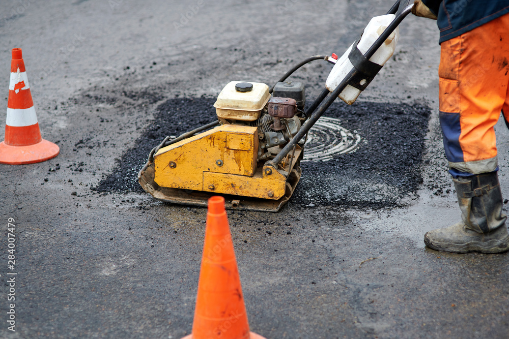 Construction worker in uniform operate vibratory plate compactor. Pothole repair process. Asphalt tamping machine operator. Pothole patching and repair. Laying asphalt around the hatch. New asphalt