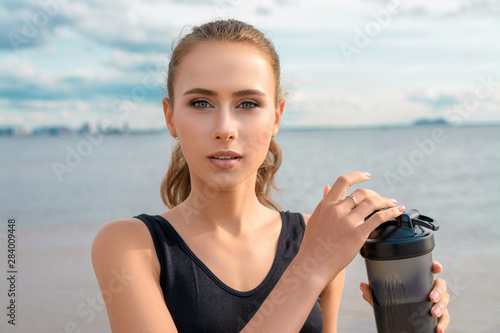 Slender athletic girl in sports clothes with a bottle of water in hands standing on the beach against the beautiful sky