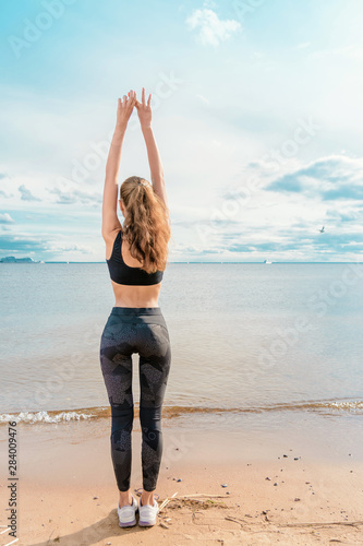 Slender athletic girl in sports clothes doing exercise on the beach against the beautiful sky