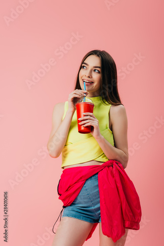 beautiful smiling girl in bright clothes drinking soda from straw isolated on pink