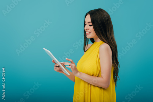 side view of smiling girl in yellow dress using digital tablet on turquoise background