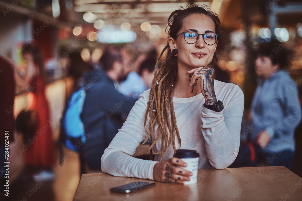 Beautiful cheerful girl with tattooes and dreadlocks is sitting at food court while drinking coffee.